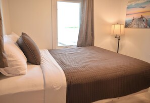 Una cama con cuidada presentación, con un edredón marrón, un almohábil blanco y otro almohadón blanco a la izquierda. Hay una ventana con cortinas entreabiertas, y una lámpara al lado derecho de la cama.