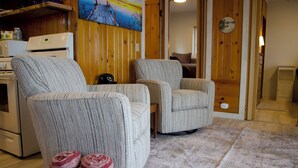 A living room with two armchairs, a sofa, a kitchen area with a microwave, and a painting on the wall.