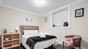 A bedroom with a wooden headboard bed, a bedside table with a lamp, a chair, a window with blinds, and a wall-mounted shelf. - Quiet 1BD in North Vancouver (North Vancouver)