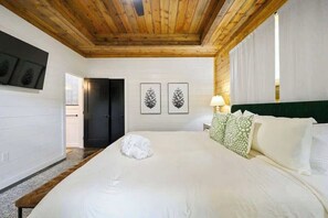 Ein Schlafzimmer mit Holzbalkendecke, weißen Wänden, einem Bett mit weißer Bettwäsche, zwei Wandbildern mit Tannenzapfen und einem grünen Kissen mit Muster.