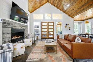 A spacious living room with a fireplace, leather sofa, patterned rug, and a high ceiling with wooden beams. - NEW! Heated Pool, Hot Tub, Fire Pit, Outdoor Lounge - Sleeps 7 (Broken Bow)