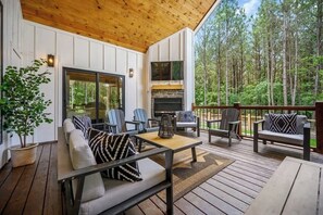 A covered outdoor seating area with a fireplace, wooden deck, and a view of a forest. - NEW! Heated Pool, Hot Tub, Fire Pit, Outdoor Lounge - Sleeps 7 (Broken Bow)