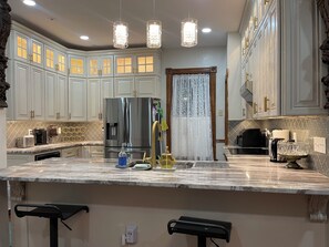 Una cocina con armarios blancos, un refrigerador de acero inoxidable, encimera de mármol e iluminación colgante.
