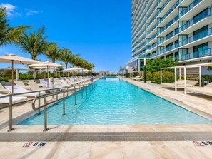 A swimming pool with lounge chairs and umbrellas, surrounded by palm trees and a modern building. - FIFA World Cup Beach Condo | Free Beach Club Access (Miami Beach)