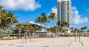 A beach with a volleyball net, palm trees, and a tall hotel building. - North Beach 1BR Condo with Balcony & Beach Club (Miami Beach)