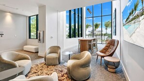 A modern hotel lobby with a large glass wall, comfortable seating, and a view of a pool and palm trees. - North Beach 1BR Condo with Balcony & Beach Club (Miami Beach)