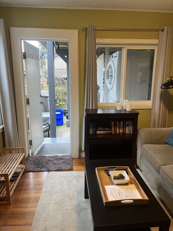 A living room with a sofa, a coffee table with a tray, a bookshelf, a window with curtains, and a door leading outside. - Snowbirds welcome to best city/climate in Canada (Victoria)