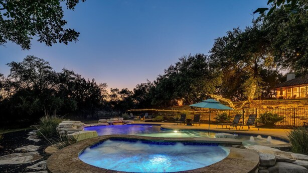 A hot tub surrounded by a stone deck, a pool, and a patio area with chairs and string lights. - Spacious retreat, pool and hot tub within walking distance of Lake Travis. (Spicewood)