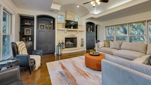 A spacious living room with a fireplace, a ceiling fan, and large windows. - Spacious retreat, pool and hot tub within walking distance of Lake Travis. (Spicewood)