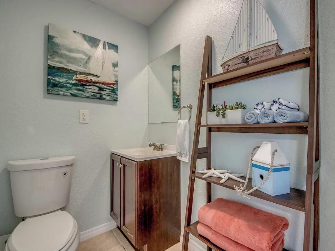 A bathroom with a toilet, sink, wooden vanity, shelf with towels, and a sailboat painting.