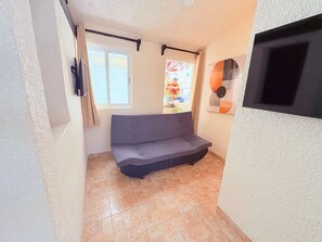 Une petite pièce avec un canapé, une fenêtre, une porte et un téléviseur fixé au mur.
