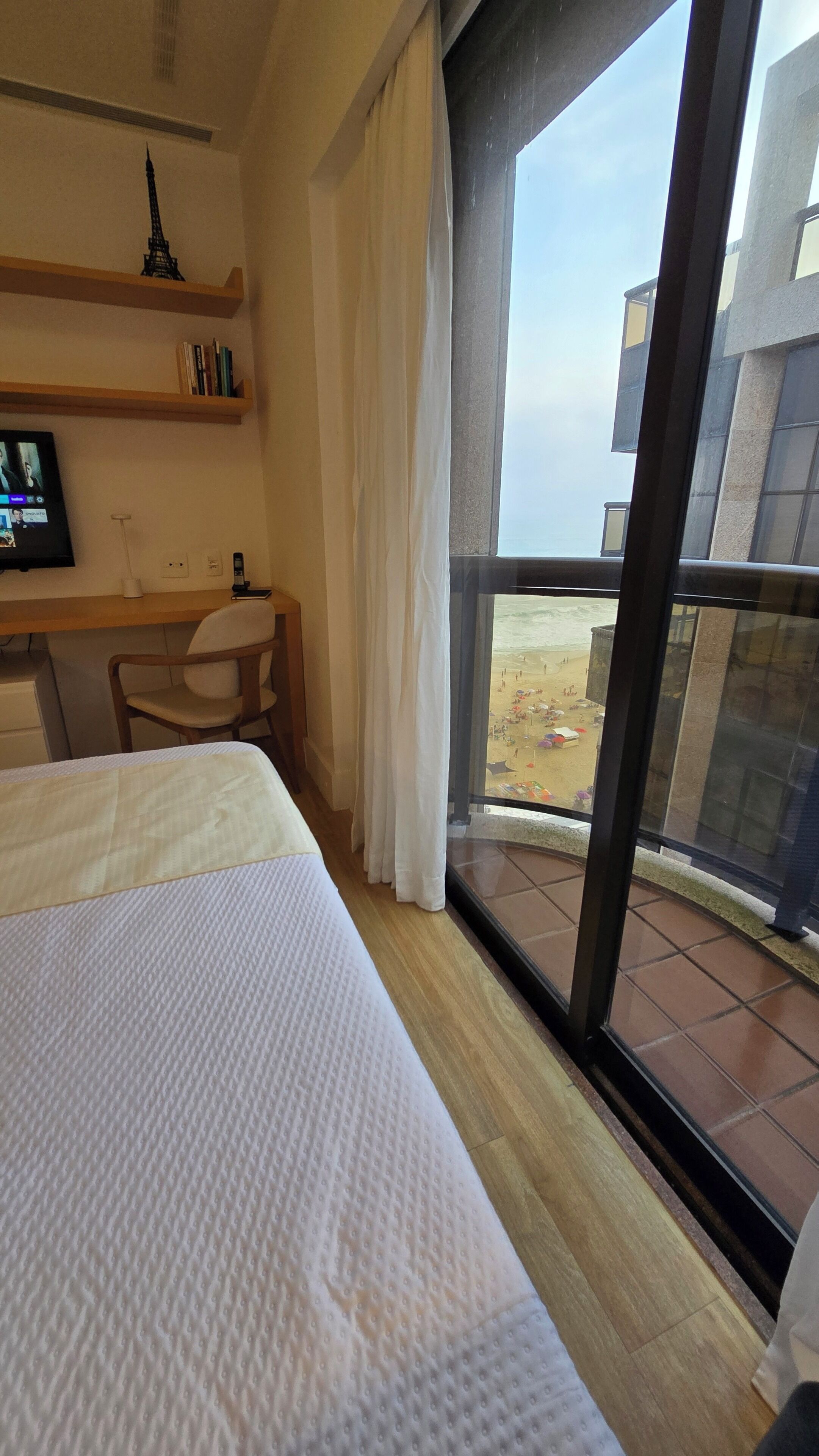 Hotelkamer met een bed, een bureau, een stoel, een televisie, een boekenkast en uitzicht op het strand door een groot raam.