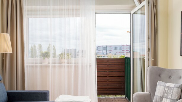 Ein Zimmer mit einer Couch, einem Sessel, einem Fenster mit durchsichtigen Vorhängen und Blick auf Gebäude.