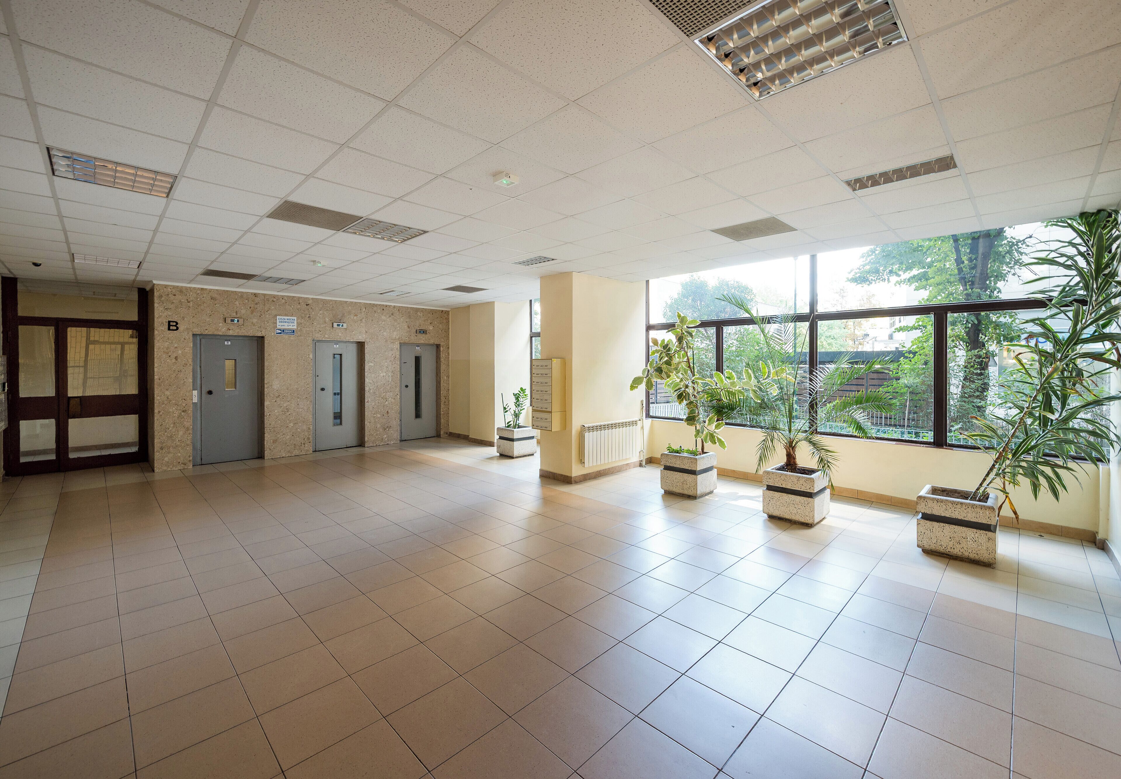 A spacious lobby with tiled floors, a reception desk, three doors, and potted plants.