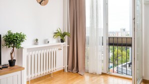 A room with a wooden floor, a white radiator, a potted plant, a clock, and a view through a window with a curtain. - Rondo ONZ Urban Core Apartment (Warszawa)
