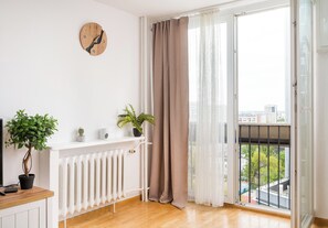 Ein Zimmer mit Holzboden, einem weißen Heizkörper, einer Topfpflanze, einer Uhr und Blick durch ein Fenster mit Vorhang.