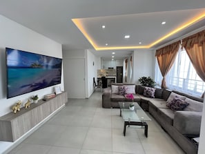 Ein modernes Wohnzimmer mit Flachbildfernseher, einer Sofagarnitur, einem Glastisch und einer Küche im Hintergrund.