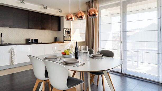A modern dining area with a round table, white chairs, and a kitchen in the background.
