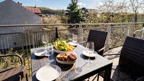 A dining table with plates, bowls of fruit, and wine glasses, set on a balcony with wicker chairs and a view of buildings and trees.