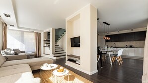 A modern living room with a sofa, a coffee table, and a TV mounted on the wall. There is a staircase leading to an upper level, and a kitchen area with a dining table and chairs.
