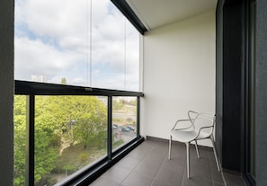 Ein Balkon mit Blick auf Bäume und einen Parkplatz, ein weißer Stuhl und ein gefliester Boden.