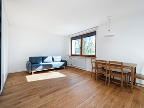 Ein Zimmer mit Holzboden, einem blauen Sofa, einem Holztisch mit Stühlen und einem Fenster mit Blick auf Bäume.