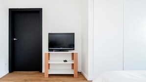 A room with a bed, a black door, a television on a white stand, and wooden flooring.