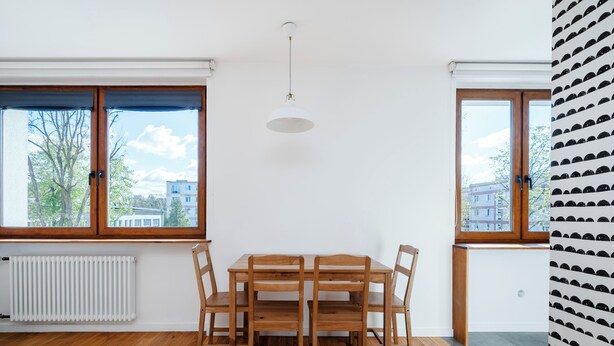 A dining area with a wooden table and chairs, large windows with blinds, a white radiator, and a ceiling light fixture.