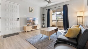 Un salon moderne doté d’un téléviseur à écran plat, d’une table basse, d’un canapé avec des coussins, d’un ventilateur de plafond et d’une porte-fenêtre coulissante donnant sur un espace extérieur.