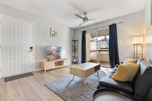 Una sala de estar moderna con televisor de pantalla plana, una mesa de centro, un sofá con cojines, un ventilador de techo y una puerta corrediza de vidrio que da a un espacio exterior.