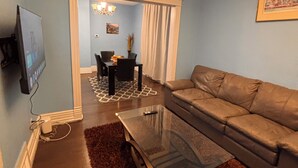A living room with a sofa, a glass coffee table, a TV mounted on the wall, a chandelier, and a dining area visible through a doorway.