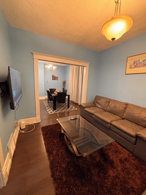 A living room with a sofa, a glass coffee table, a TV mounted on the wall, a chandelier, and a dining area visible through a doorway. - Two story House: 3 Bed room 1/5 Bathroom (Winnipeg)