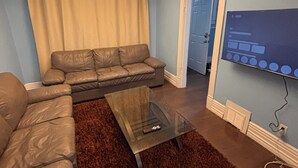 A living room with a sectional sofa, a glass coffee table, a brown rug, a ceiling light, and a TV mounted on the wall.