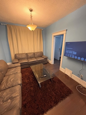 A living room with a sectional sofa, a glass coffee table, a brown rug, a ceiling light, and a TV mounted on the wall. - Two story House: 3 Bed room 1/5 Bathroom (Winnipeg)