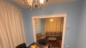 A dining area with a black table, chairs, and a chandelier. There is a living room in the background with a sofa and a coffee table.