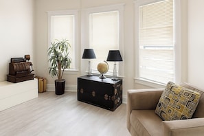 A living room with a light wooden floor, a beige sofa with patterned cushions, a small black side table with a globe and lamps, a potted plant, and a vintage typewriter on a white dresser. - Detroit 4BR Home | Bathtub and Pet Friendly Stay (Detroit)
