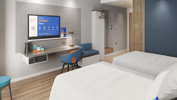 A modern hotel room with a bed, a desk with a computer, a chair, a TV mounted on the wall, and a wardrobe.