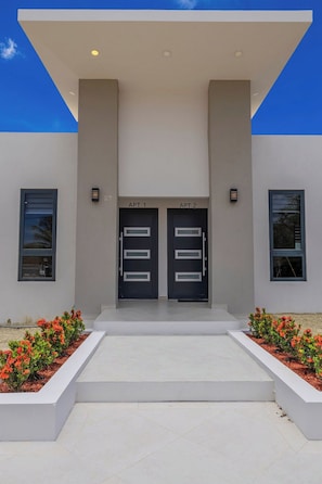 A modern building with two doors labeled 'APT 1' and 'APT 2' at the entrance, flanked by windows and surrounded by a landscaped area with red flowers. - BRAND NEW 3BR2BA @ Palm Beach w/ Shared Pool & BBQ (Noord)