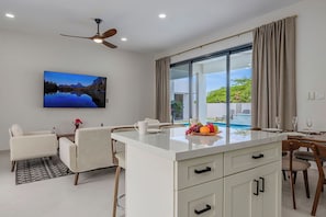 A modern kitchen with a central island, dining area with chairs, a ceiling fan, and a large window with curtains. - BRAND NEW 3BR2BA @ Palm Beach w/ Shared Pool & BBQ (Noord)