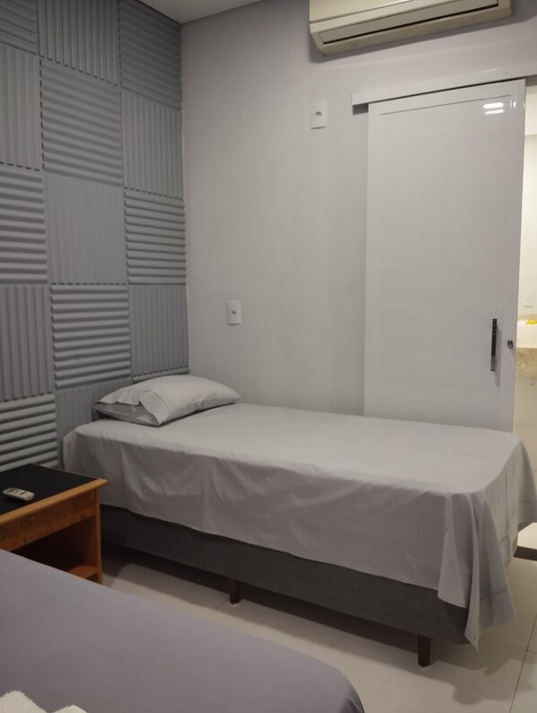 Un lit simple avec un matelas gris et du linge de lit blanc, une table de chevet avec une télécommande, une armoire à porte coulissante et un climatiseur fixé au mur.