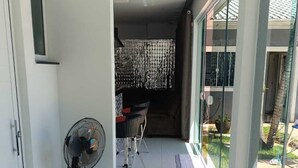 Ein Flur mit einem Ventilator, ein Essbereich mit Stühlen und einem Tisch sowie ein Blick ins Freie durch große Fenster.