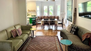 Ein Wohnzimmer mit einer Couch, einem Couchtisch, einem Esstisch mit Stühlen, einem Deckenventilator und großen Fenstern.