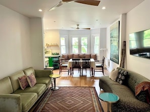 Ein Wohnzimmer mit einer Couch, einem Couchtisch, einem Esstisch mit Stühlen, einem Deckenventilator und großen Fenstern.