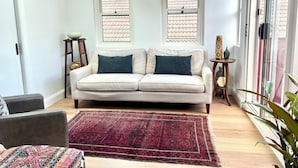 Ein Wohnzimmer mit einer Couch, einem gemusterten Teppich, zwei Fenstern und einem kleinen Tisch mit einer Dekoration.
