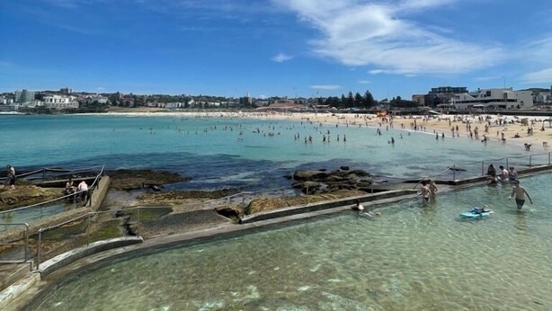 Eine Küstenlandschaft mit Strand, klarem Wasser, felsigen Bereichen und einer fernen Uferlinie mit Gebäuden.