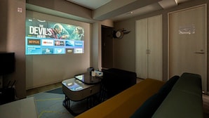 Et hotelværelse med et stort tv, der viser en film, et skrivebord med stol, en sofa og et klædeskab.