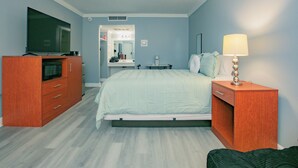 Quarto com cama, televisão, mesa de cabeceira com luminária, cômoda e espelho.
