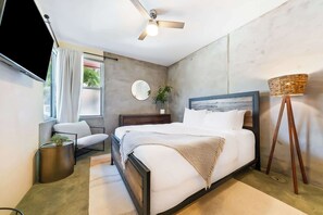 Ein modernes Schlafzimmer mit Bett, Deckenventilator, Fernseher, Nachttisch mit Lampe und Fenster mit Vorhängen.