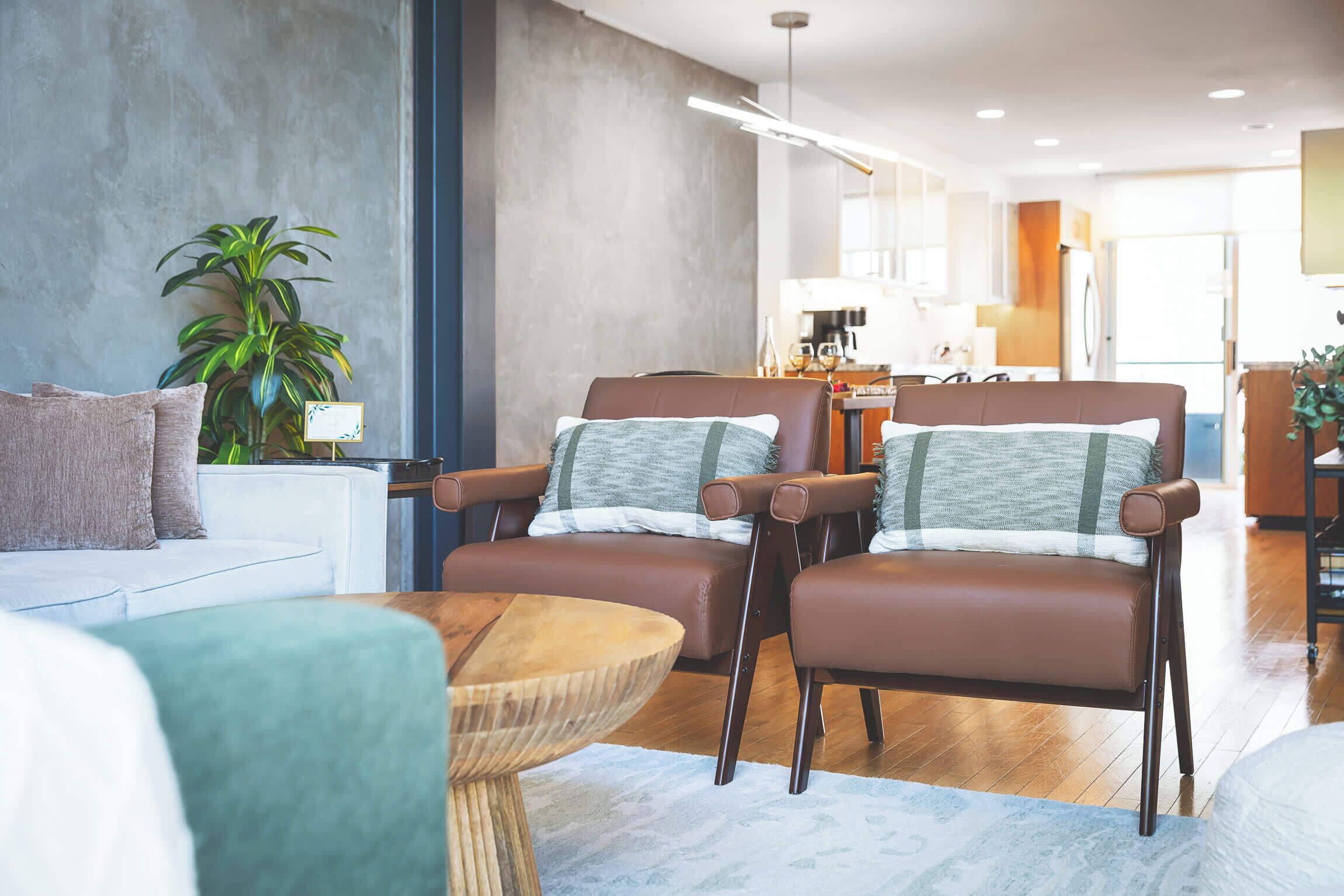 Ein modernes Wohnzimmer mit einer Couch, zwei braunen Ledersesseln, einem runden Holztisch und einem Blumentopf.
