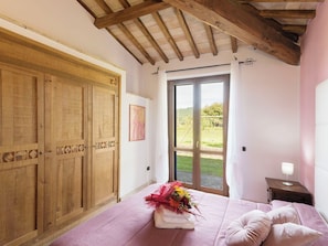 Ein Schlafzimmer mit einem Holzschrank, einem Bett mit rosa Bettdecke, einem Nachttisch mit Lampe und einer Glastür zu einem Garten.